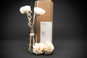 Sample Palo Santo Reed Diffuser set displayed on a black backdrop.  Kraft box labelled with product scent.  Jar next to box displays how the set look.  Jar includes two sola flowers, two curly reed stick and three reed sticks in the jar. Lying in between the jar and box are two other sola flowers.