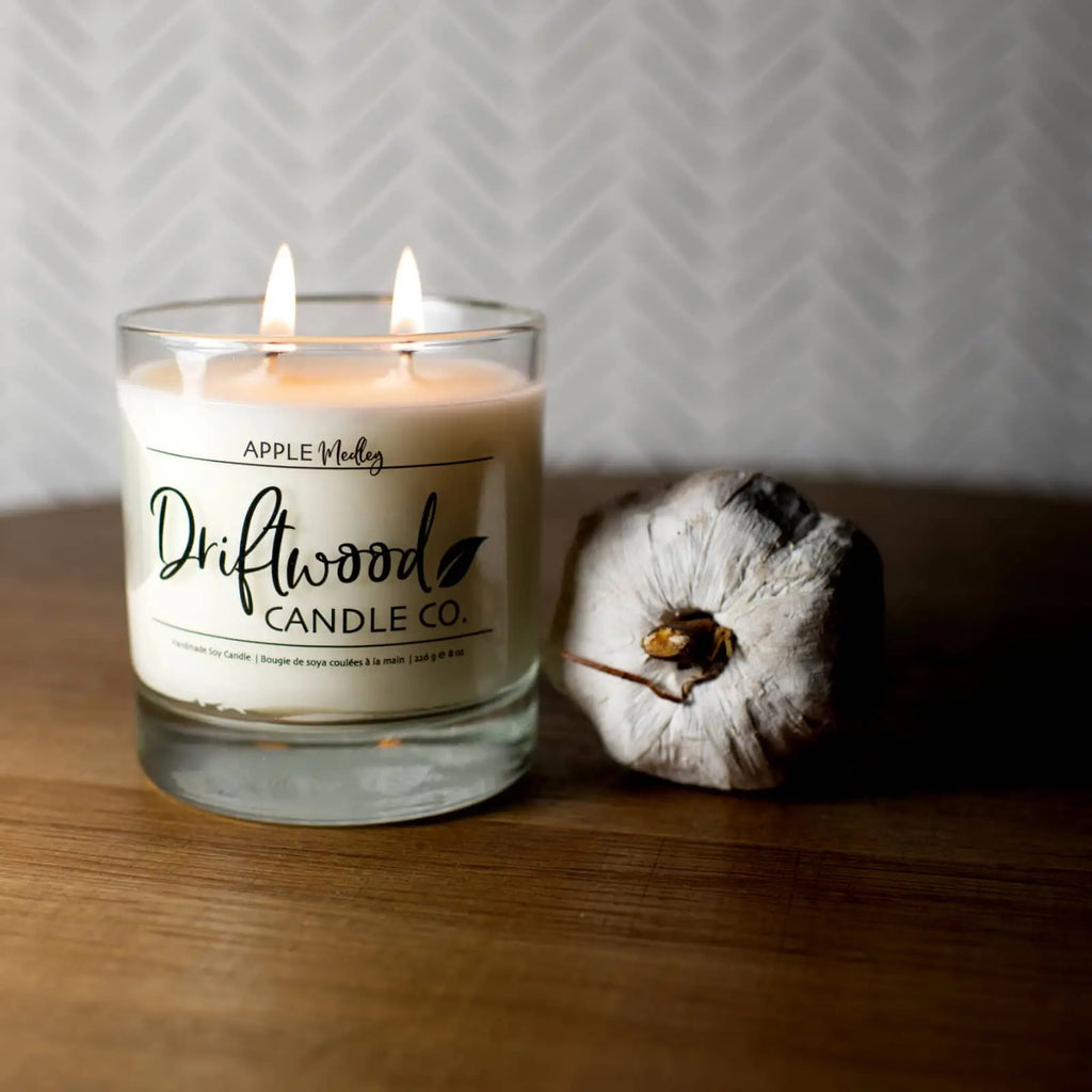 Driftwood Candle Co. apple medley scented candle sitting on a wood table with a decorative wood apple next to it.  The reusable whiskey glass has the scent label affixed to it.  Both double wicks are lit to show the gentle glow of the candle.